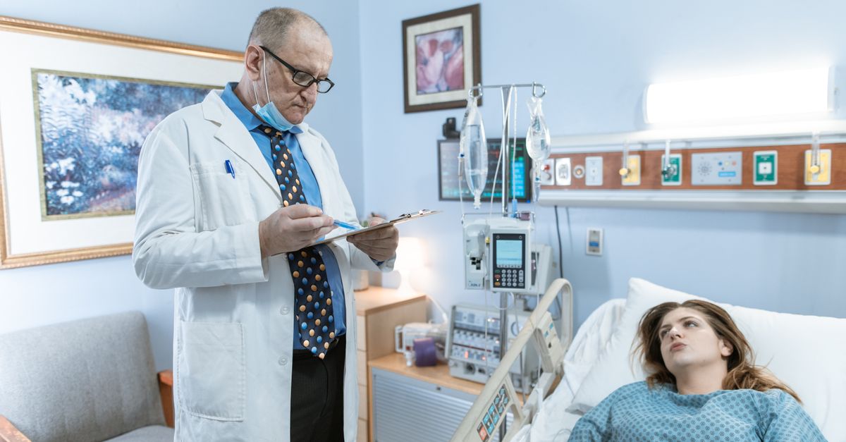 Doctor in white coat checking patient chart in hospital room with medical equipment.