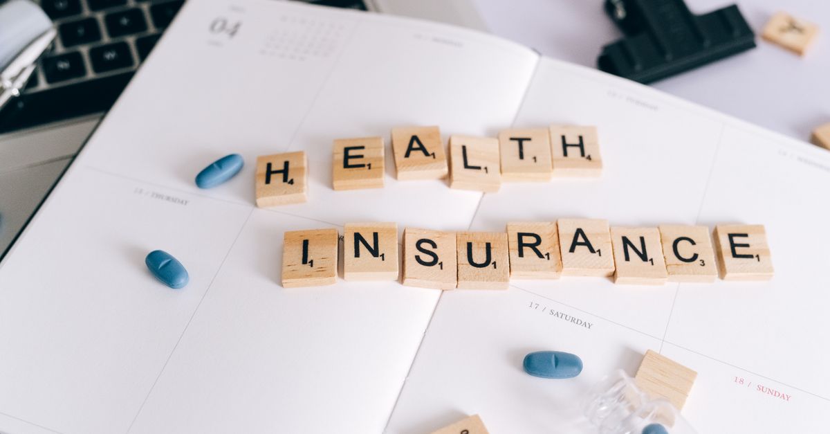 Scrabble tiles spelling 'Health Insurance' on planner with pills and laptop, symbolizing healthcare planning.