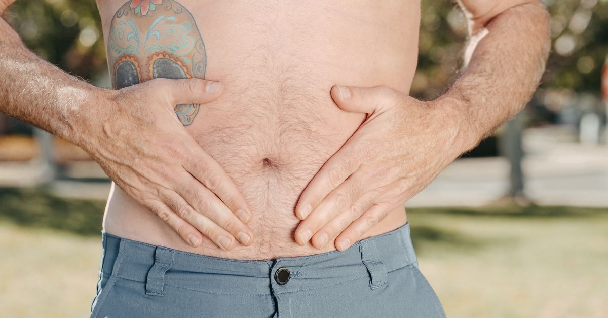 A man with a tattoo on his torso holds his stomach outdoors, suggesting discomfort.